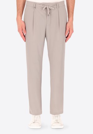 Uomo con pantaloni beige su misura con pieghe e coulisse in vita, abbinati a sneakers bianche con lacci, in piedi con le braccia lungo i fianchi.