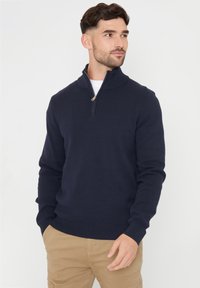 Námořnicky modrý svetr se zapínáním na zip a stojáčkem. Vyrobený z měkké pleteniny, má stojatý límec a žebrované manžety. Kombinován s khaki kalhotami. Jednoduchý, klasický design.