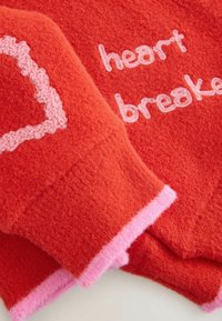 Rode fleece stof met een roze hartmotief en geborduurde tekst "heartbreaker"; beschikt over een zachte textuur en contrasterende roze bies.