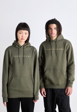 Dos jóvenes adultos vistiendo sudaderas oliva a juego con el texto "TOMMY JEANS", de pie uno al lado del otro contra un fondo claro y liso.