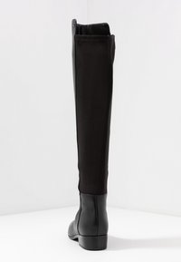 Bota negra por encima de la rodilla con un eje de tela elegante y un tobillo de cuero. Punta redondeada y tacón bajo apilado para mayor estabilidad. Detalles de costura presentes.