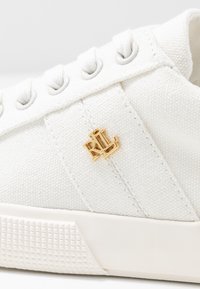 Primer plano de un zapato de lona blanco con cordones blancos y el logotipo dorado "RLL" en el lateral, encima de una suela de goma texturizada blanca.