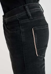 Jean en denim noir avec une coupe ajustée, comportant une poche latérale accentuée d'une rayure multicolore. Tissu texturé et taille standard.