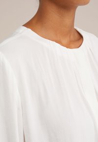 Nærbillede af en person iført et hvidt tøj med en draperet halsudskæring mod en ensfarvet beige baggrund.