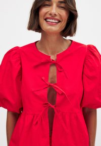 Rode blouse met pofmouwen, voorzien van een ronde halslijn, strikhangertjes aan de voorkant en een uitlopende onderkant met een gestructureerde stofafwerking.
