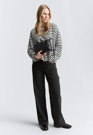 Borsa a spalla nera con hardware circolare; indossata con un maglione oversized a righe bianche e nere e jeans neri a vita alta e a gamba larga.