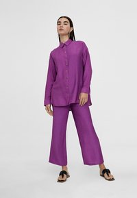 Camisa de botones morada con cuello, combinada con pantalones morados de corte suelto. La tela parece texturizada. El modelo lleva sandalias con un detalle de lazo.