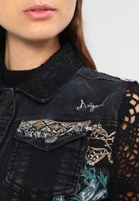 Černá džínová vesta s vyšívanými vzory, krajkovým límcem a dekorativními akcenty ve stříbrné, zlaté a tyrkysové barvě. Má třepící se detaily a kapsy.