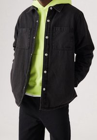 Černá džínová bunda se zapínáním na knoflíky, dvěma kapsami na prsou a mírně oversized střihem. Nositelná přes výrazně zelenou mikinu s kapucí.
