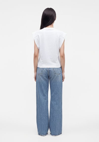 Personne avec de longs cheveux raides et foncés, de dos, portant un haut blanc sans manches et un jean bleu ample dans un studio simple.