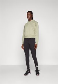 Hoodie cropped verde-azeitona com cordões, leggings pretas ajustadas e sapatilhas de desporto pretas com detalhes em branco no logo.