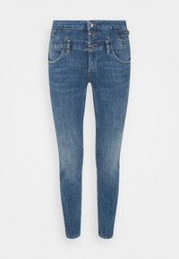 Jeans ajustados de color azul intermedio con cierre de triple botón, bolsillos frontales y trabillas para cinturón, mostrando un ligero desgaste en los muslos y las rodillas.