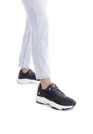 Zapatillas atléticas negras con suelas blancas, acentos texturizados y detalles en dorado en los cordones. Combinadas con unos vaqueros rectos de lavado claro.