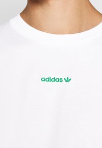 Vit bomull t-shirt med grönt broderat "adidas"-logotyp och Trefoil-mönster på bröstet; rund halsringning; mjuk textur.