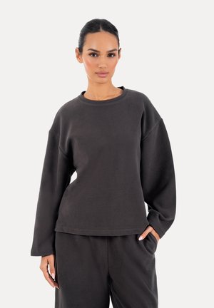 Kvinde stående med hånden i lommen iført mørkegrå langærmet sweatshirt og matchende bukser på lys baggrund.