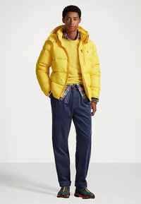 Gul puffjacka med huva, lager-på-lager över en gul tröja och en rutig skjorta, kombinerad med marinblå manchesterbyxor och flercoulrad sportskor.