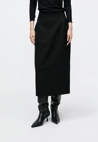 Čierna sukňa po kolená z hladkej látky s priliehavým pásom a jemnými švovými detailmi, kombinovaná s elegantnými čiernymi čižmami na podpätku.