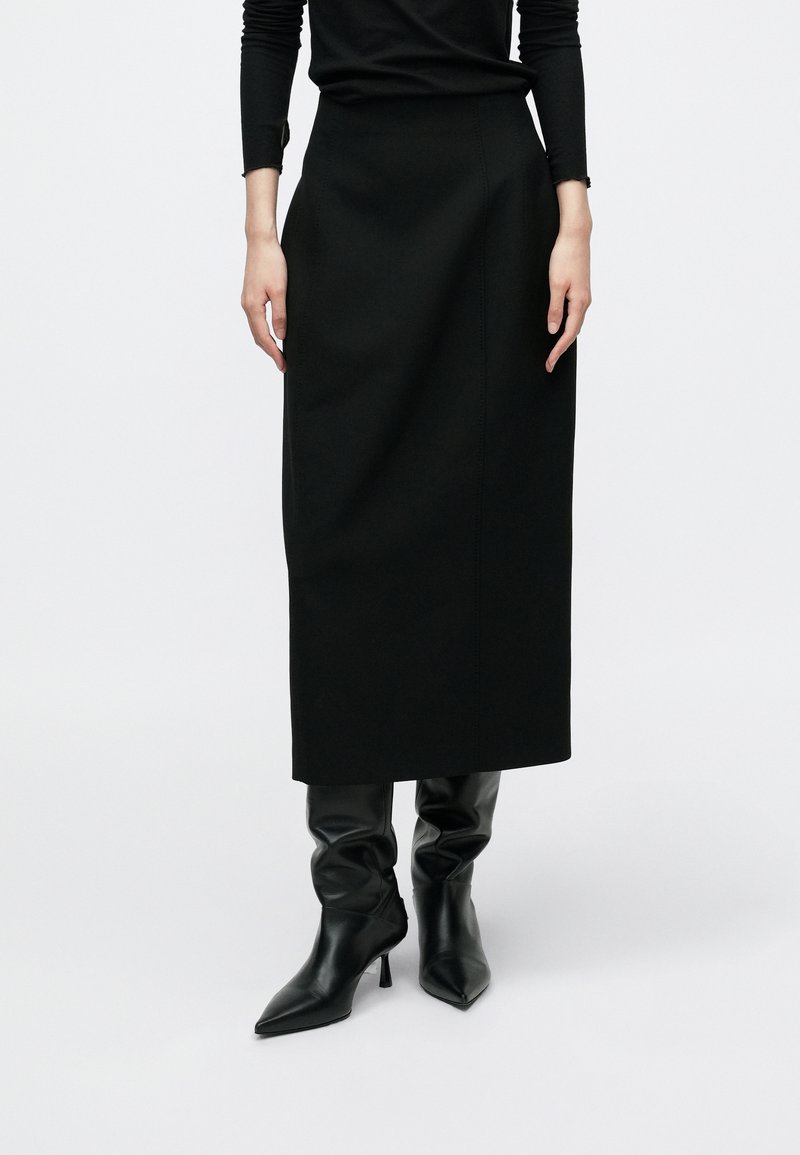 Čierna sukňa po kolená z hladkej látky s priliehavým pásom a jemnými švovými detailmi, kombinovaná s elegantnými čiernymi čižmami na podpätku.