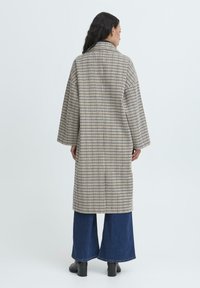 Donna con lunghi capelli scuri che indossa un cappotto a quadri beige e blu navy, jeans larghi blu e stivaletti neri con tacco, in piedi di spalle.