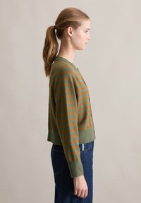 Jonge vrouw in profiel met lichtbruine paardenstaart, gekleed in een groen en bruin gestreept vest en een blauwe spijkerbroek, tegen een effen achtergrond.