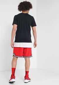 Černé tričko s krátkým rukávem přes bílý síťovaný top na ramínka, doplněné červenými šortkami s černo-bílým lemováním a červenými ponožkami se vzory.