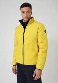Giacca a vento gialla con zip e colletto neri, caratterizzata da un logo circolare sulla manica sinistra. Tessuto morbido e design aderente.