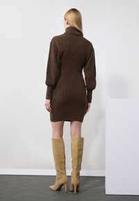 Brun ribbestrikket kjole med høy krage og figursydd midje, kombinert med knelange beige semskede støvler, vist fra bak.