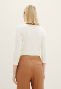 Kobieta z blond włosami do ramion, ubrana w białą bluzkę z długim rękawem i brązowe spodnie z wysokim stanem, stojąca na jednolitym tle.