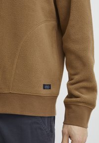 Sudadera de forro polar marrón con una superficie texturizada, puños acanalados y una pequeña etiqueta negra cerca del dobladillo. Cuenta con un bolsillo tipo canguro.