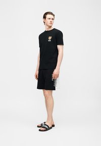 Νέος άνδρας που φοράει μαύρο t-shirt Moschino με λογότυπο αρκουδάκι, μαύρο σορτς με πλαϊνό κείμενο και μαύρα slides με λευκή επωνυμία.
