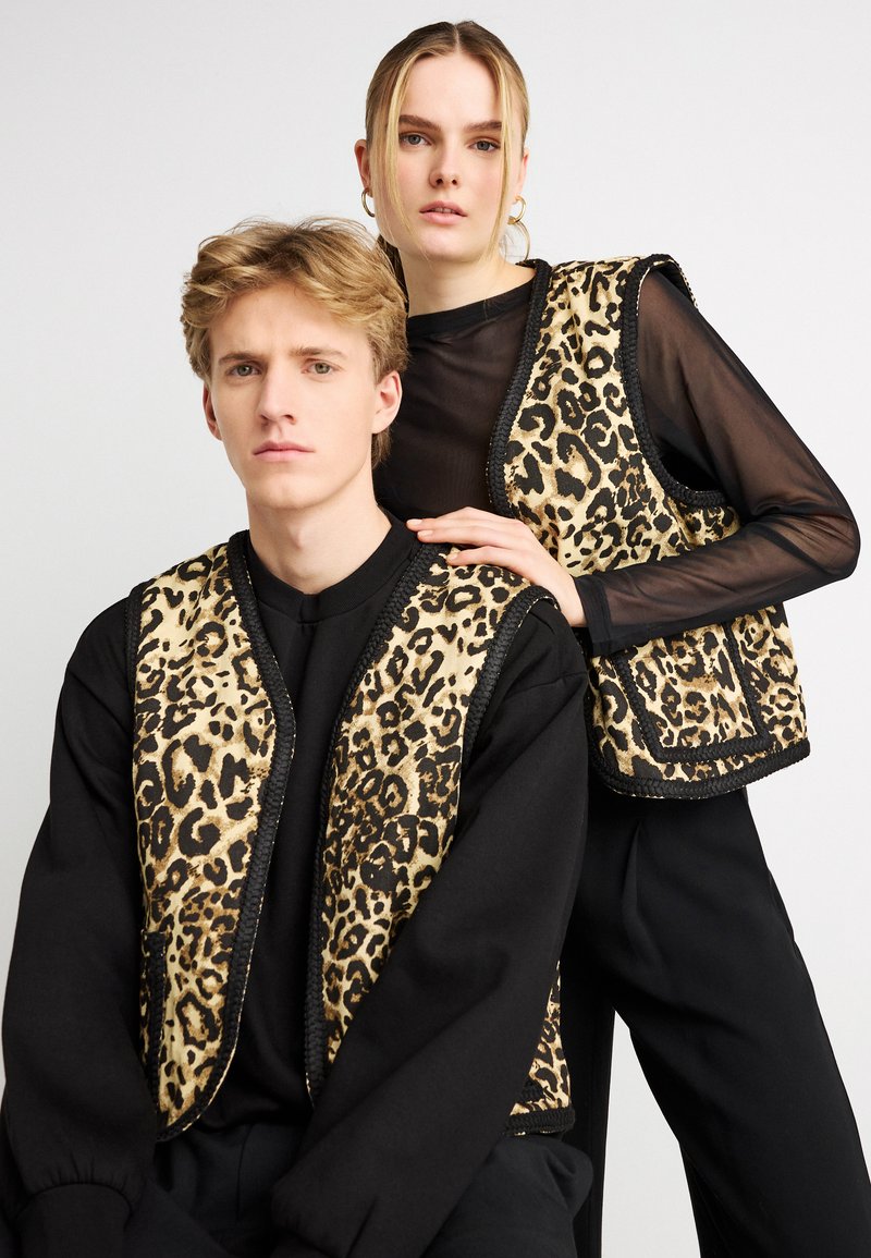 Barna és fekete anyagból készült leopárdmintás mellények, kontrasztos fekete szegéllyel, fekete felsők fölött viselve. A pár közel áll egymáshoz semleges pózban.