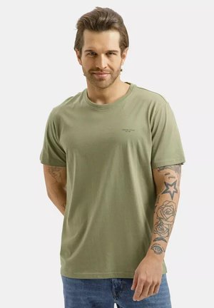 Mężczyzna z jasnobrązowymi włosami, ubrany w zieloną koszulkę i niebieskie dżinsy, pokazujący tatuaże, w tym gwiazdę, róże i oko na prawym przedramieniu.