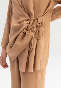 Camicia in tessuto leggero color marrone con un lato arricciato e Legature, patta con bottoni e dettagli plissettati per texture e struttura.