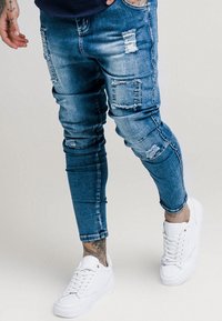 Úzké modré džíny z denimu s obnošenými záplatami a třepenými okraji, v kombinaci s bílými teniskami. Hladká textura se středním odstínem.