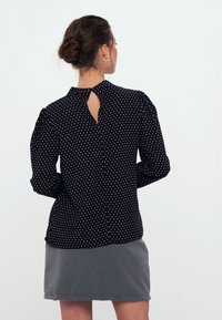 Schwarze Bluse mit Polka-Dots, langen Ärmeln, gerafften Schultern und einem Schlitzverschluss am Rücken. Das Material wirkt leicht und strukturiert.