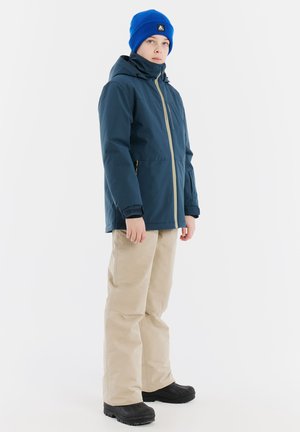 Marineblaue wasserdichte Jacke mit Reißverschluss, beige Schneehosen und schwarzen Stiefeln. Die Jacke hat eine Kapuze und eine blaue Strickmütze.