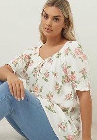 Vit blommig blus med rosa rosor och gröna löv. Lätt passform, rynkad halsringning, korta puffärmar. I kombination med blå denimjeans.
