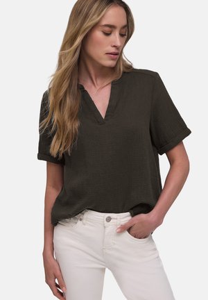 Kobieta ubrana w ciemnozieloną bluzkę z krótkim rękawem i dekoltem w serek oraz białe spodnie, z lewą ręką w kieszeni, patrząca w dół w lewą stronę.