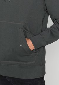 Felpa con cappuccio in cotone grigio con tasca kangaroo frontale, polsini e orlo a costine, con dettagli di cuciture visibili. Texture morbida.