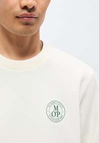 Νεαρό άτομο που φοράει ανοιχτόχρωμο φούτερ με πράσινο κυκλικό λογότυπο που γράφει "Marc O'Polo Stockholm 1967" στο στήθος.