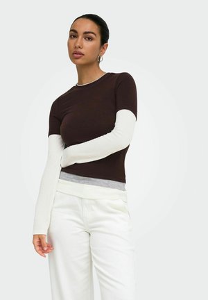 Femme aux cheveux foncés porte un haut marron à manches courtes par-dessus une chemise blanche à manches longues et un pantalon blanc, debout devant un fond uni.