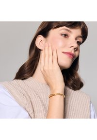 Złota bransoletka o skręconej fakturze noszona na nadgarstku. Modelka ma na sobie lekką dzianinową bluzkę, która podkreśla bransoletkę na neutralnym tle.