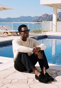 Homme portant des lunettes de soleil, un pull blanc et un pantalon noir, assis en tailleur près d'une piscine avec des montagnes et la mer en arrière-plan.