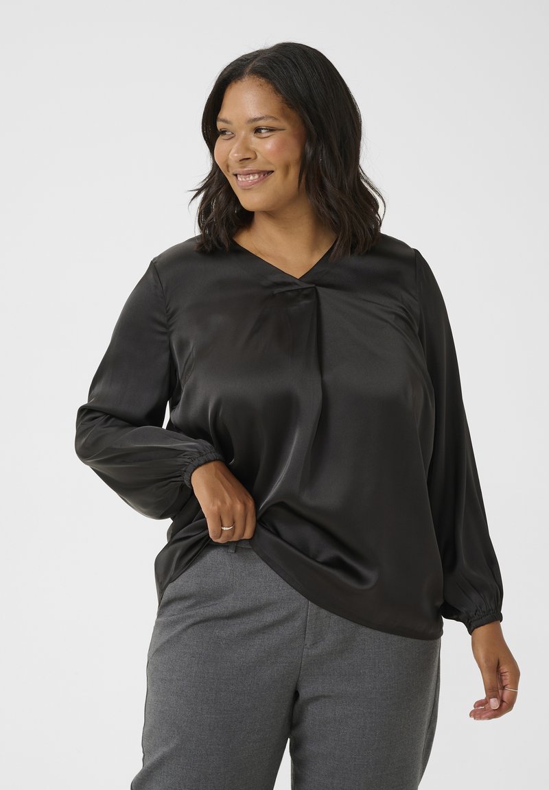 Blouse en satin noir avec un décolleté en V et des manches froncées, associée à un pantalon gris ajusté. La texture lisse et le léger éclat sont mis en avant.