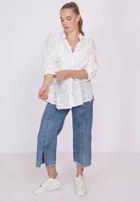 Weiße Bluse mit Blumenmuster, Puffärmeln, Knopfleiste und Rüschenausschnitt, kombiniert mit blauen, knöchellangen Jeans und weißen Sneakers.