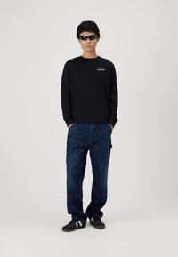 Zwarte crew-neck sweatshirt met de tekst "Club1990". Losvallende donkerblauwe jeans met voorzakken. Zwarte sneakers en slanke zonnebril.
