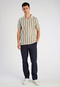 Krótki rękaw, beżowa koszulka polo w czarne pionowe paski, z klasycznym kołnierzykiem. Noszona z granatowymi spodniami i białymi butami sportowymi.