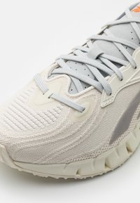 Light beige athletic shoe featuring a textured knit upper, gray laces, rubber sole with ridged accents, and subtle gray design elements.