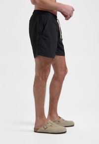 Schwarze Badeshorts mit elastischem Bund und Kordelzug, ausgestattet mit Seitentaschen. Tragen mit beigen Veloursleder-Schlüpfschuhen.