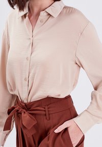 Chemise beige clair à boutons avec un col et des manches amples, associée à un pantalon taille haute de couleur rouille avec une ceinture nouée.